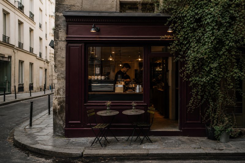 Narrow Marais street with a small café shopfront, pavement tables and morning light on old stone facades