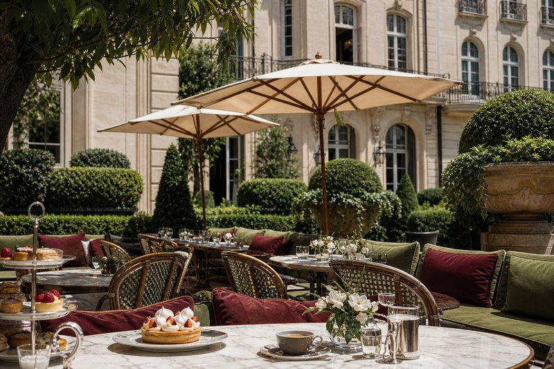 Elegant branded café terrace on a Paris luxury shopping avenue with sculpted hedges and monogrammed parasols