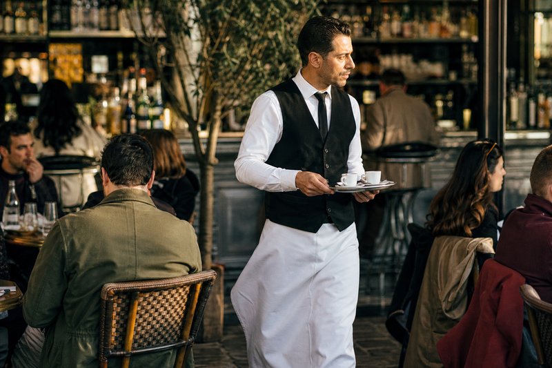Waiter in a long apron carrying espresso cups across a busy Paris café floor with a zinc counter behind