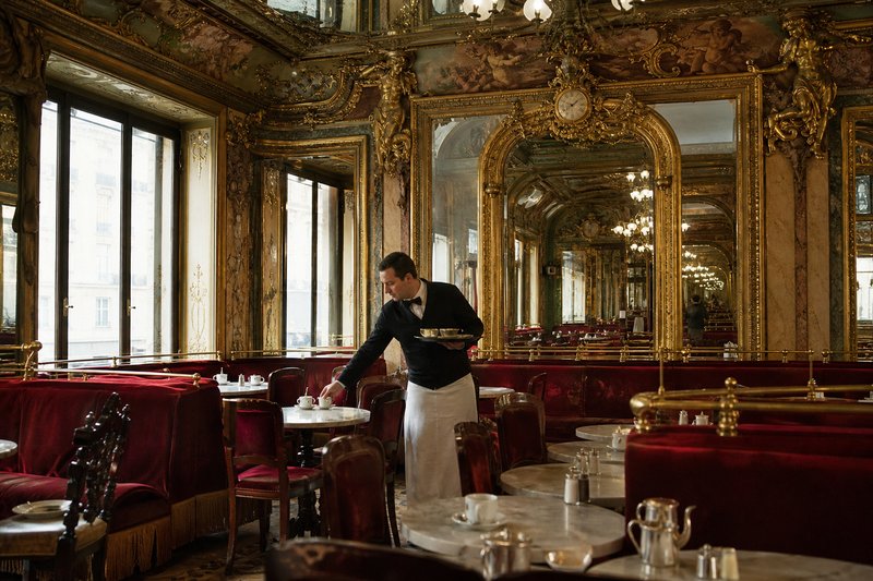 Ornate historic Paris café interior with gilded mirrors, red banquettes and a chandelier above marble tables