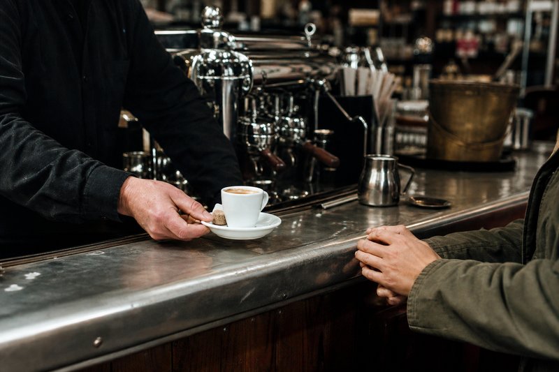 Café counter in Paris with a waiter at the espresso machine and a chalkboard listing coffee prices for counter and terrace